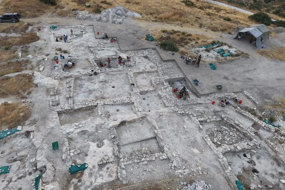 Aerial view of an excavated hilltop with a grid of low stone walls forming rectangular rooms on pale bedrock, round holes cut into the floors, and excavators at work with buckets and wheelbarrows
