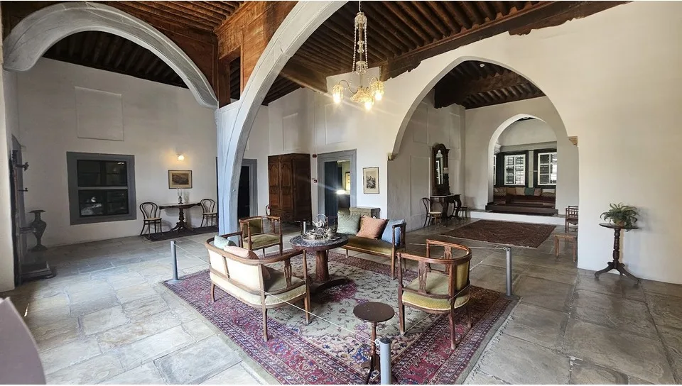 A whitewashed hall with stone arches, a dark timber ceiling and a flagstone floor, furnished with upholstered chairs, a sofa and a round table on a patterned rug, with a raised seating area visible through an arch at the back
