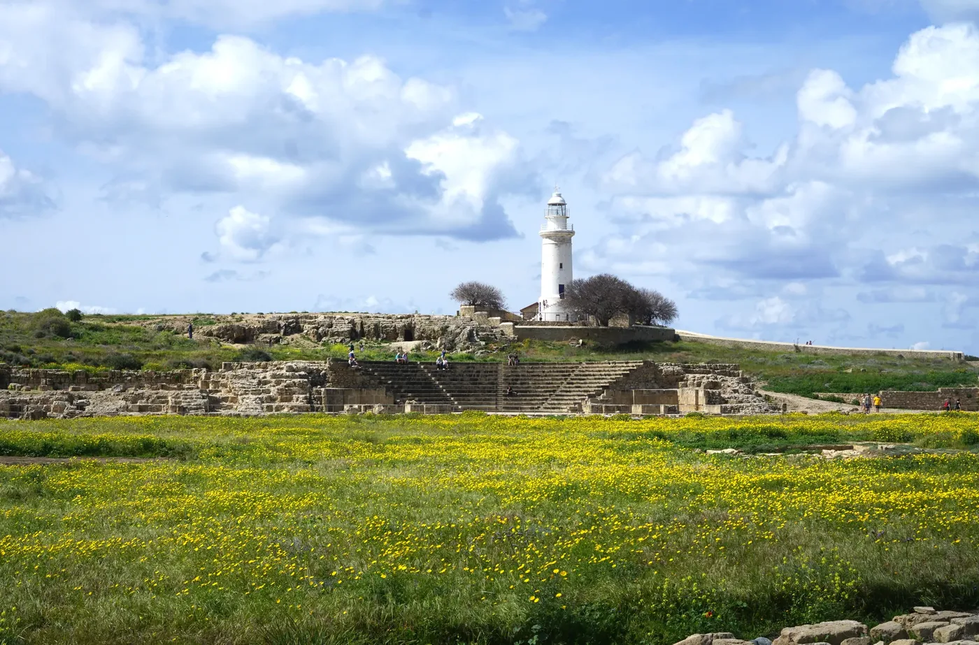 A white lighthouse on a rocky hill above the stepped stone seating of an ancient odeion, with a meadow of yellow flowers in front