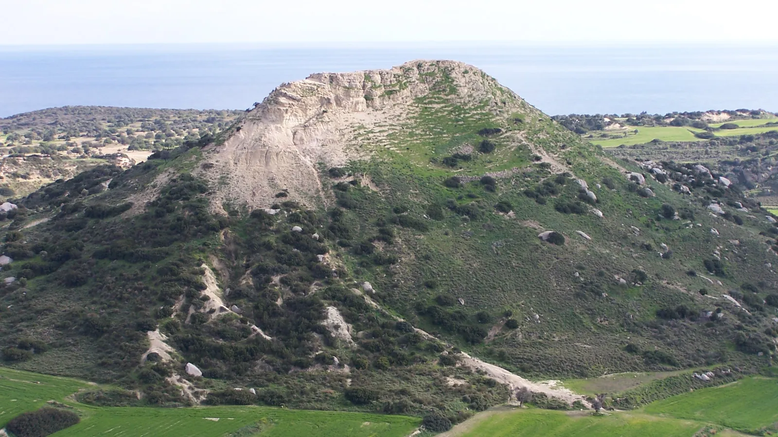 A steep, pale limestone hill with scrub-covered slopes rising above green fields, with the sea on the horizon behind it