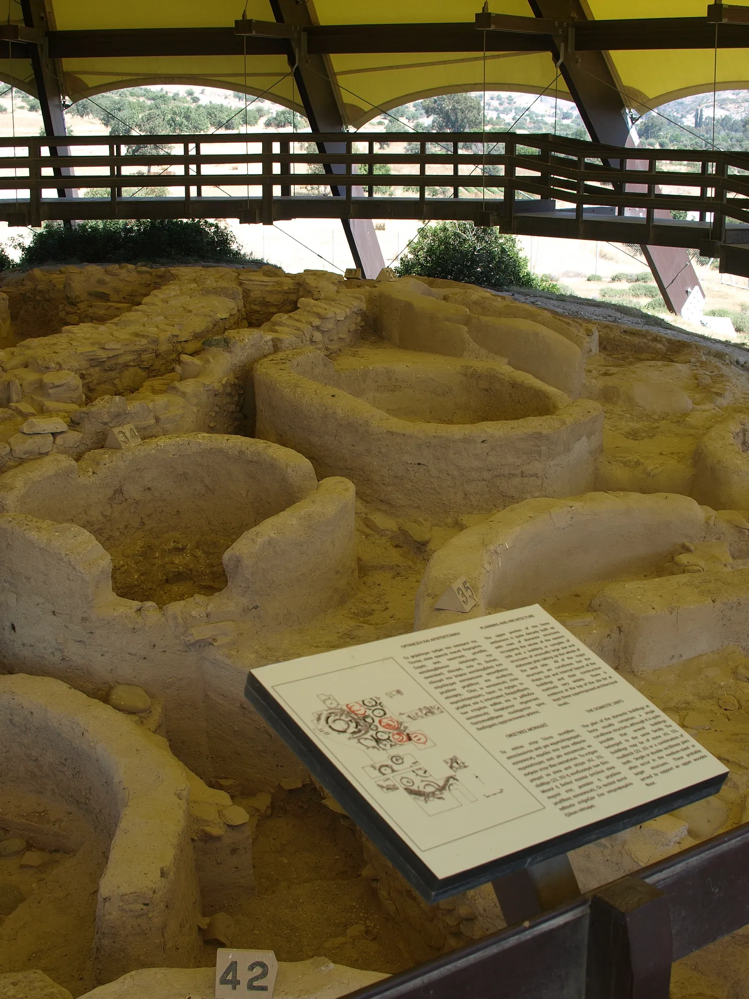 Excavated round buildings with low curved walls in yellow earth, an information panel in front and the shelter roof above
