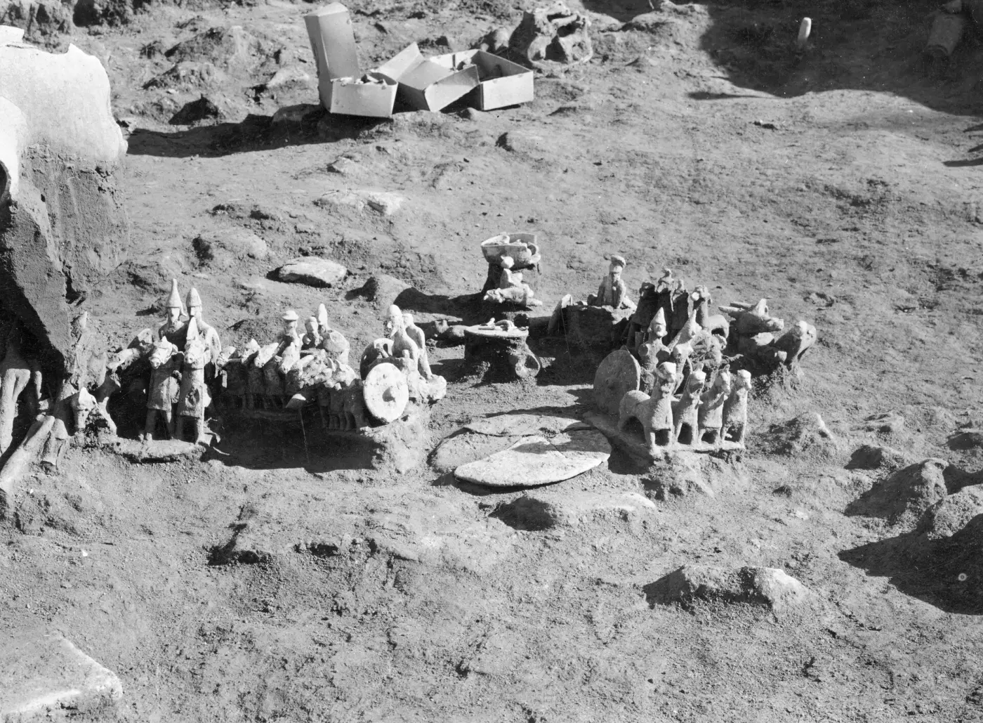 Black-and-white photograph of terracotta chariots and standing figurines lying in groups on the floor of an excavation trench, with cardboard boxes at the back