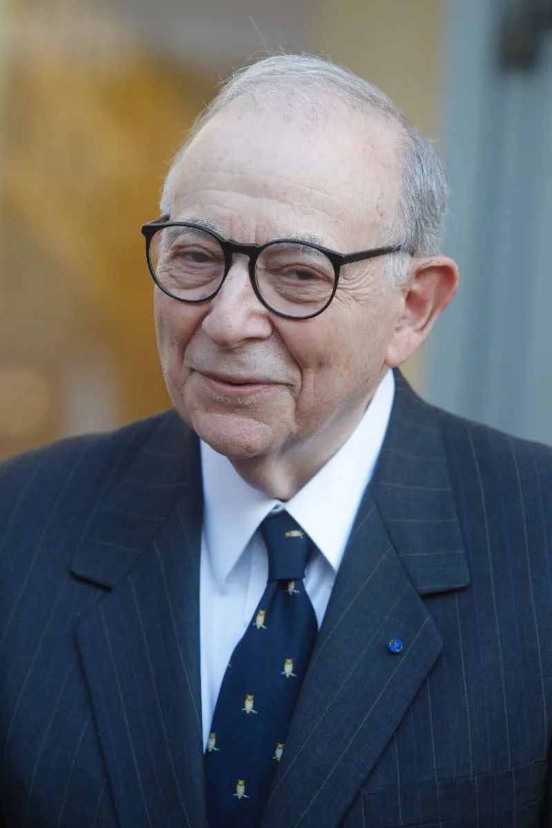 Portrait of an elderly man with white hair and round dark glasses, in a dark pinstripe suit and patterned navy tie