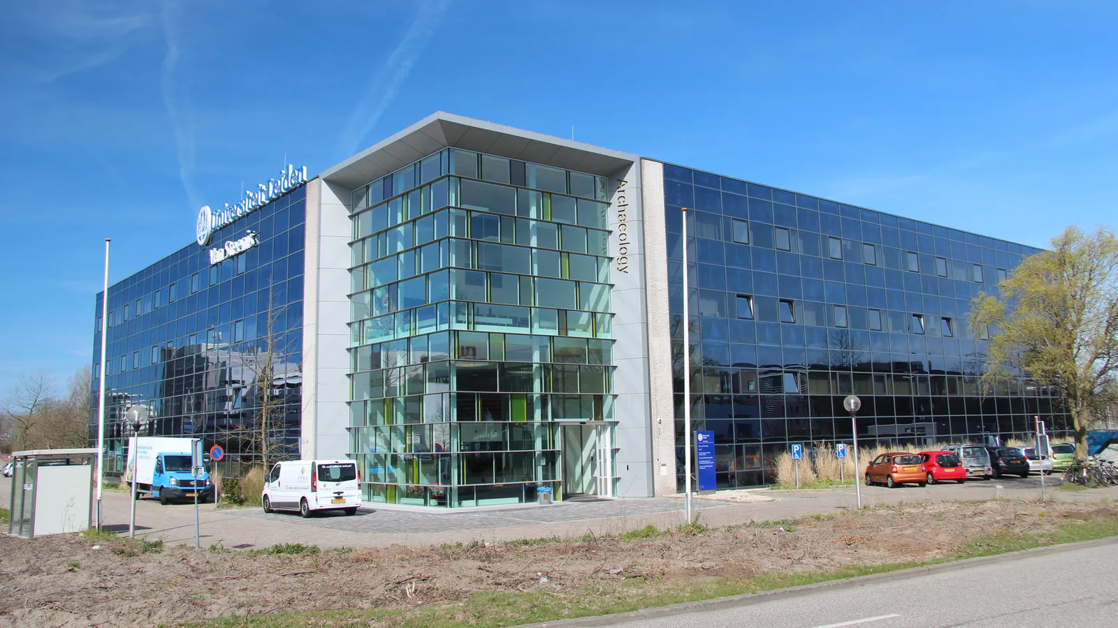 A four-storey office building clad in blue glass, with a green-tinted glass corner tower bearing the word Archaeology, vans and cars parked in front under a clear sky