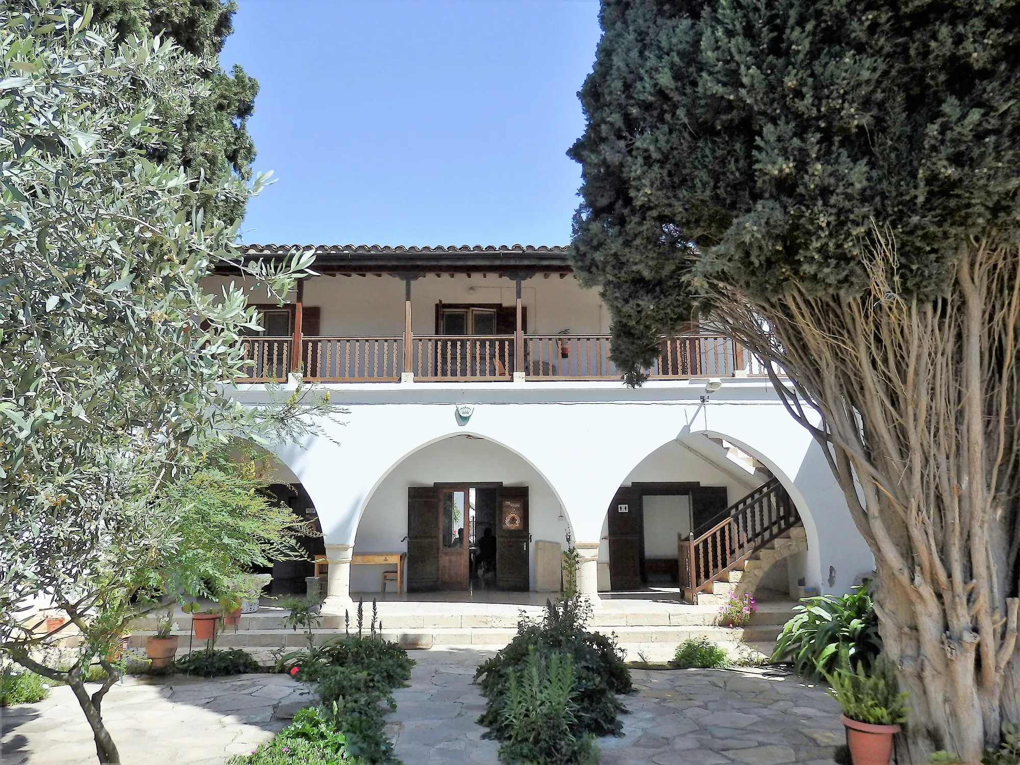 A whitewashed two-storey village house with an arched arcade and a wooden balcony, behind an olive tree and a cypress