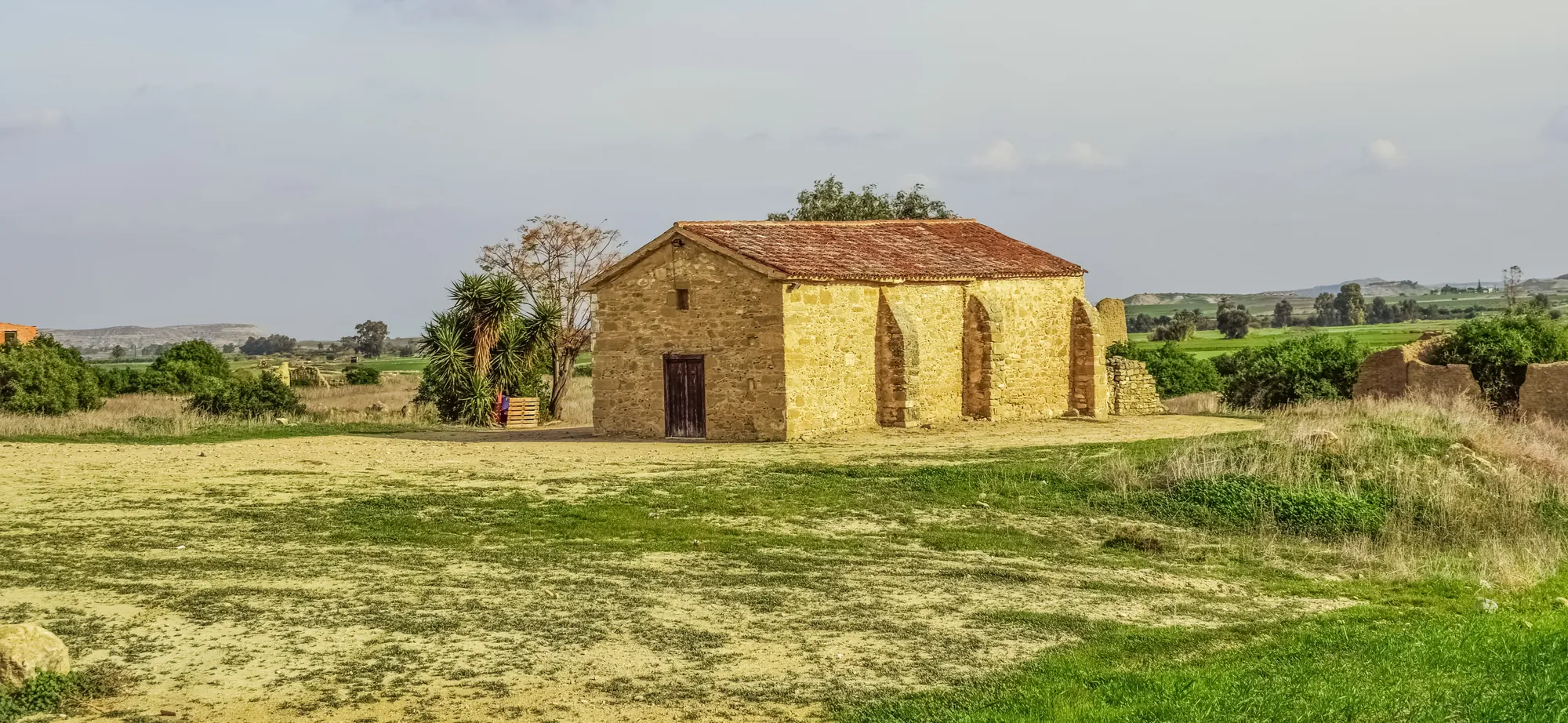 Small stone church with a red tiled roof in open grassland, ruined houses beside it, low flat-topped hills in the distance