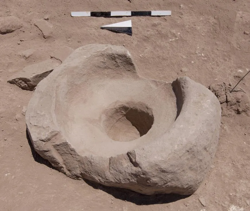 A large pale limestone block with a deep round hollow and a second shallower hollow, lying in reddish excavated earth, with a scale bar and north arrow