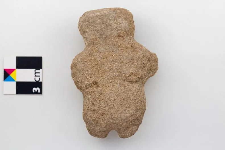 A small sandy-brown stone figure with a head, short arms and legs, shaped in outline only, beside a 3 cm scale