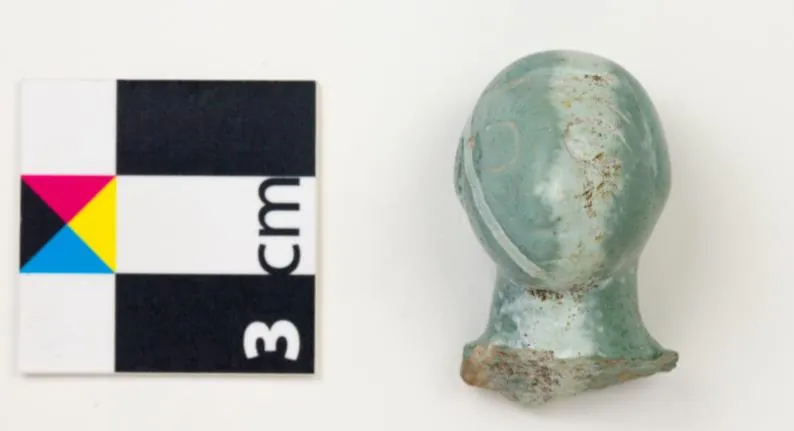 A small pale green stone head on a short neck, with a faint incised eye, beside a 3 cm scale