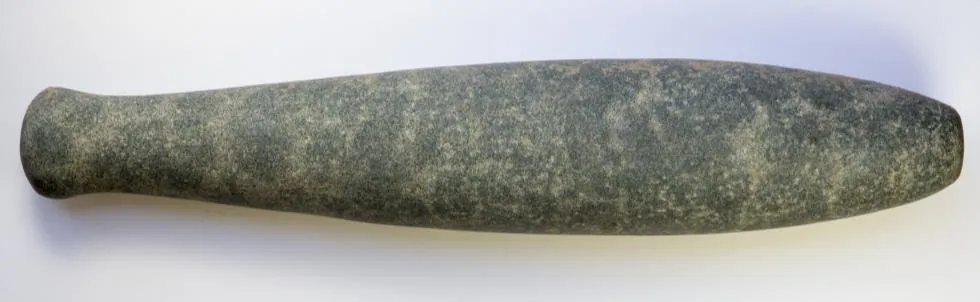 A long dark green stone pestle, thicker at one end and with a knob at the other, lying on white