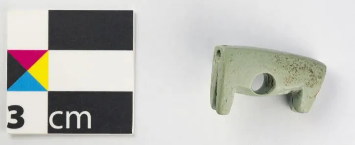 A small pale green stone animal body with four short legs and a hole through the middle, beside a 3 cm scale