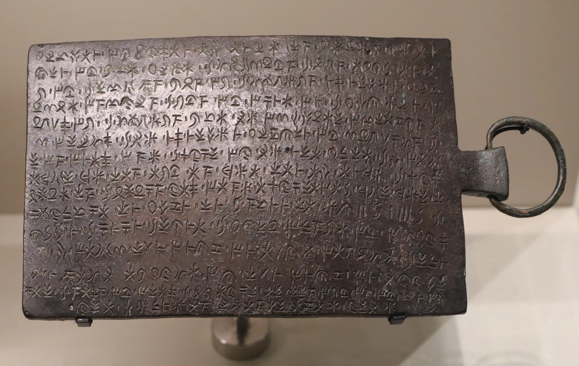 A dark rectangular bronze tablet with a ring handle, covered with lines of Cypriot syllabic signs