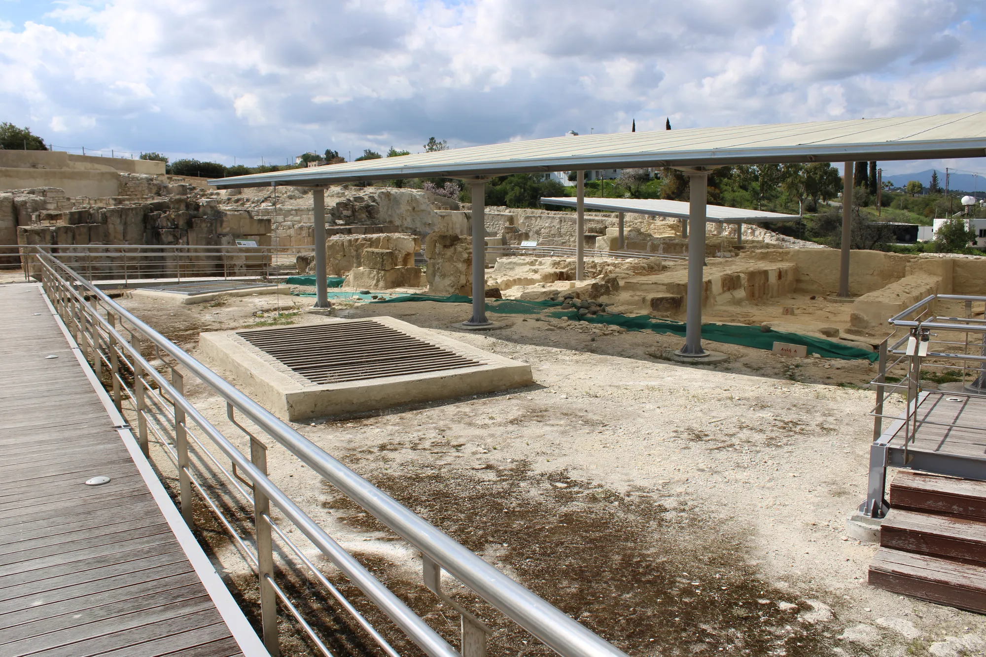 A paved courtyard with a covered well, a wooden walkway and excavated rooms under a roof