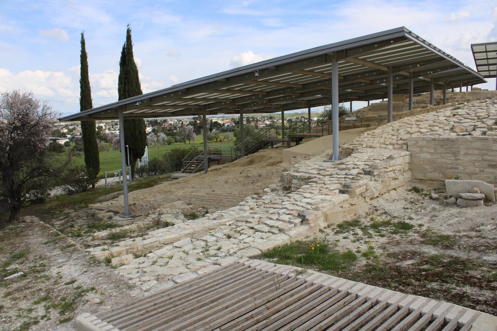 A stepped stone ramp and gateway beneath a modern shelter, with cypress trees beside it