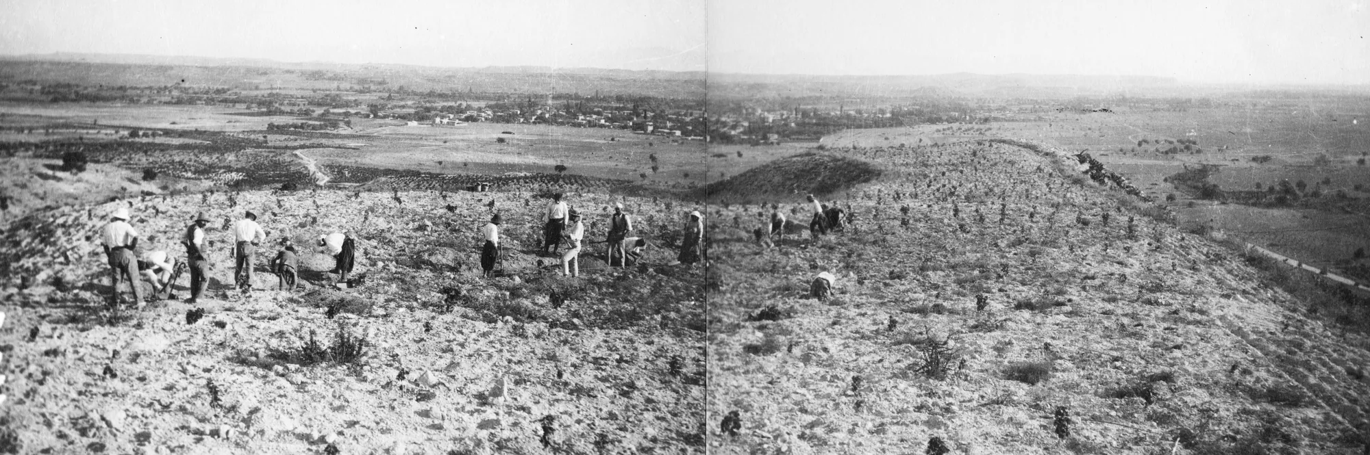 A black-and-white panorama of a bare hill with a line of workmen on the ridge, and a wide plain beyond