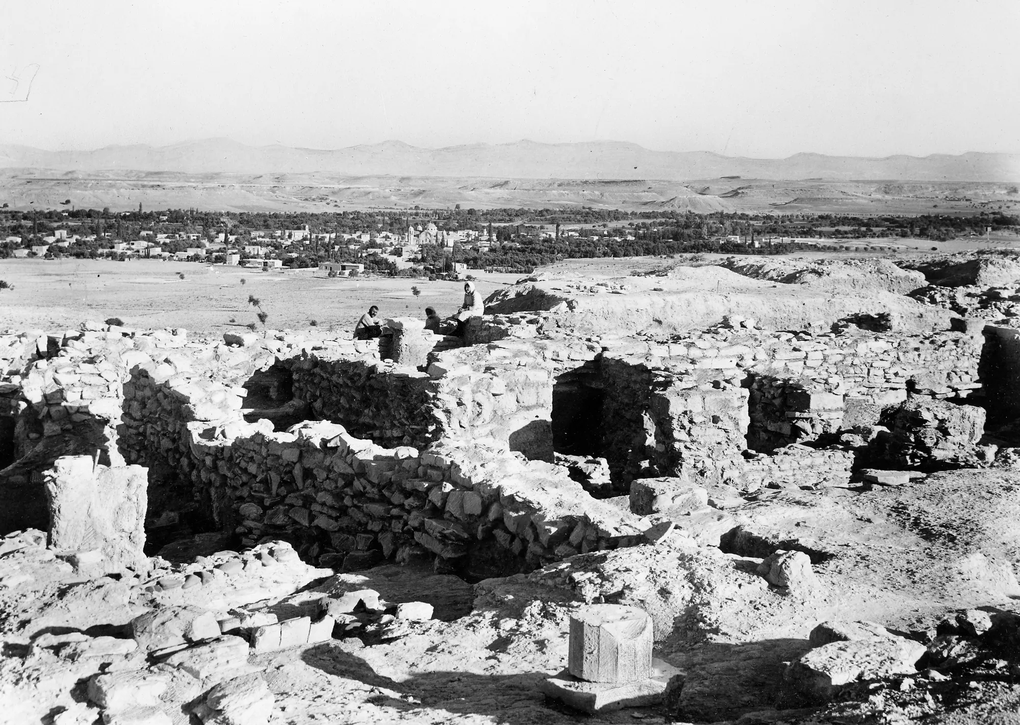 An old black-and-white photograph of excavated stone walls and rubble on a hill, with a plain and hills behind