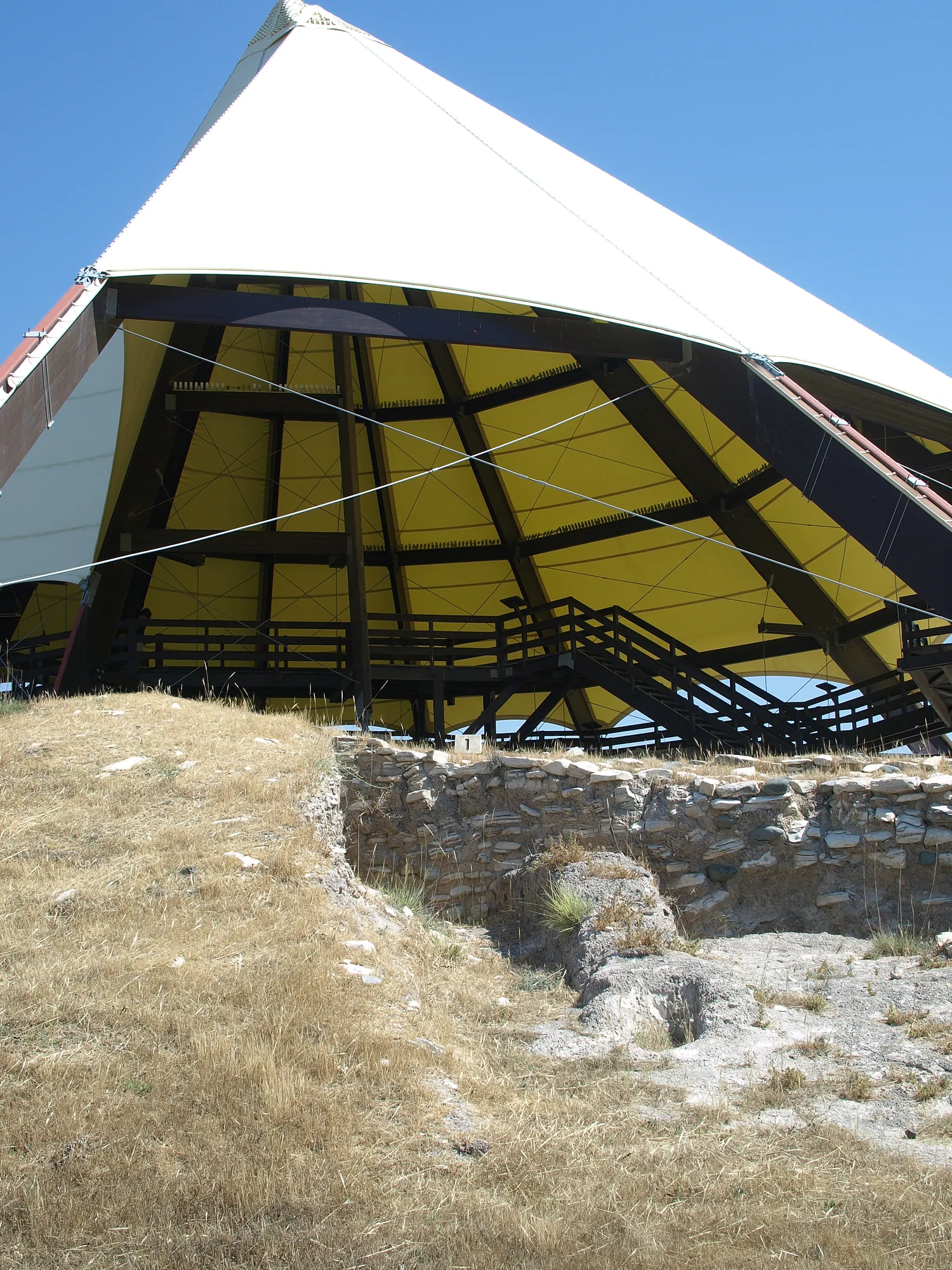 A dry grassy slope with a rough stone wall exposed in a cut, and the yellow-lined shelter roof and walkway above