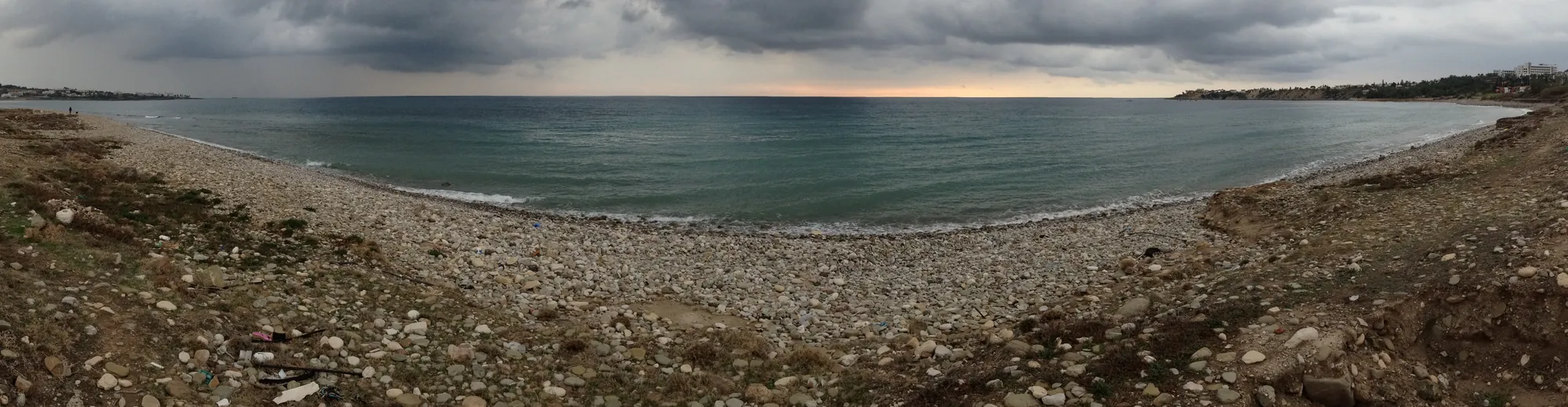A wide curving pebble beach and grey sea under heavy cloud, with low scrubby ground in the foreground