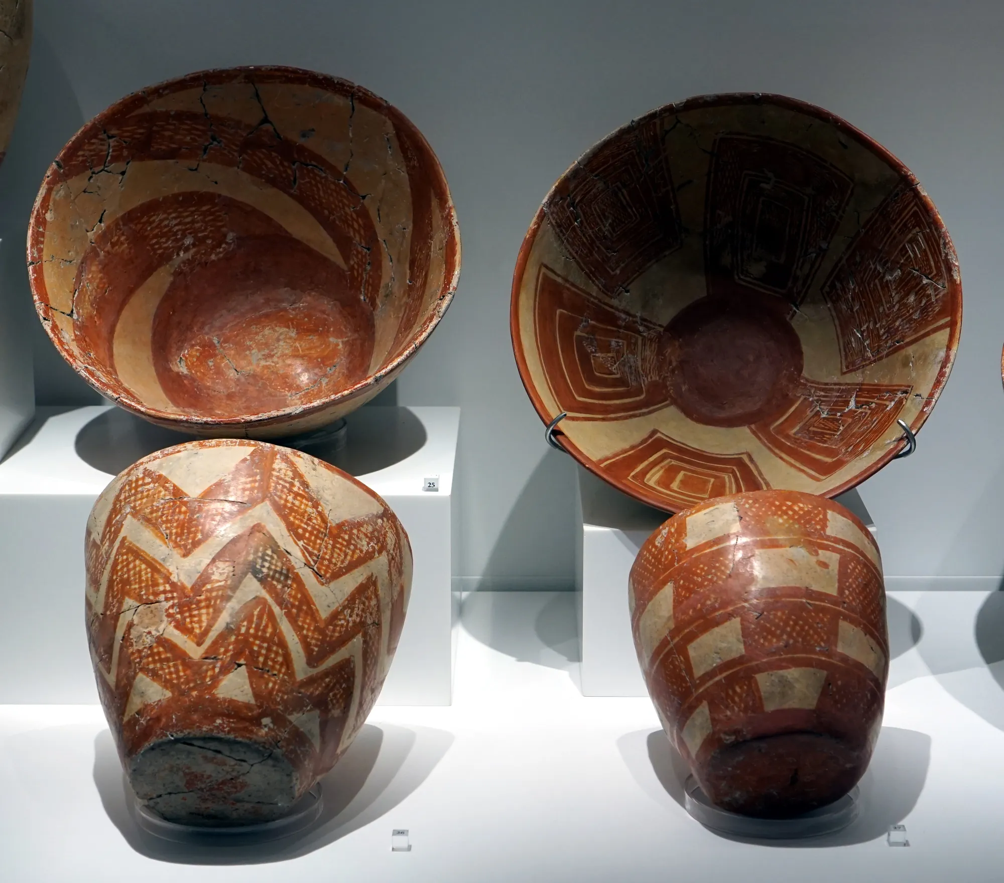 Four hemispherical clay bowls painted in red on a pale ground with lattice, zigzag and stepped designs, on museum stands