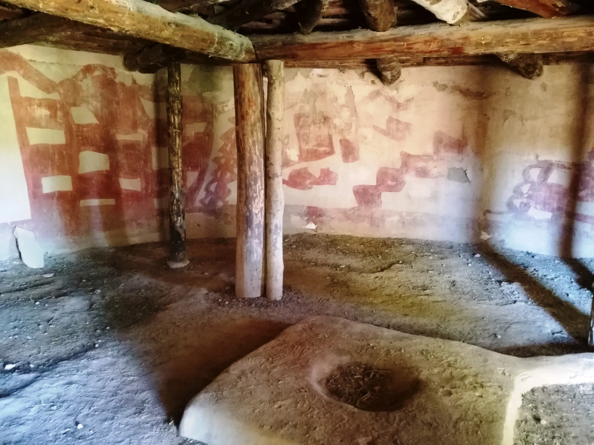 Interior of a round house with timber posts and beams, red painted wall and a large stone with a cup-shaped hollow in the floor