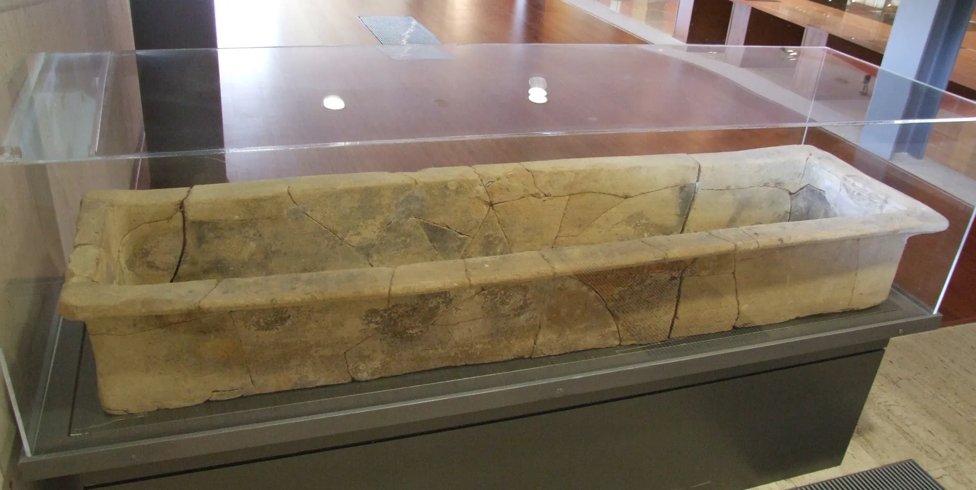 A long, shallow, rectangular clay coffin with mended cracks in a glass display case