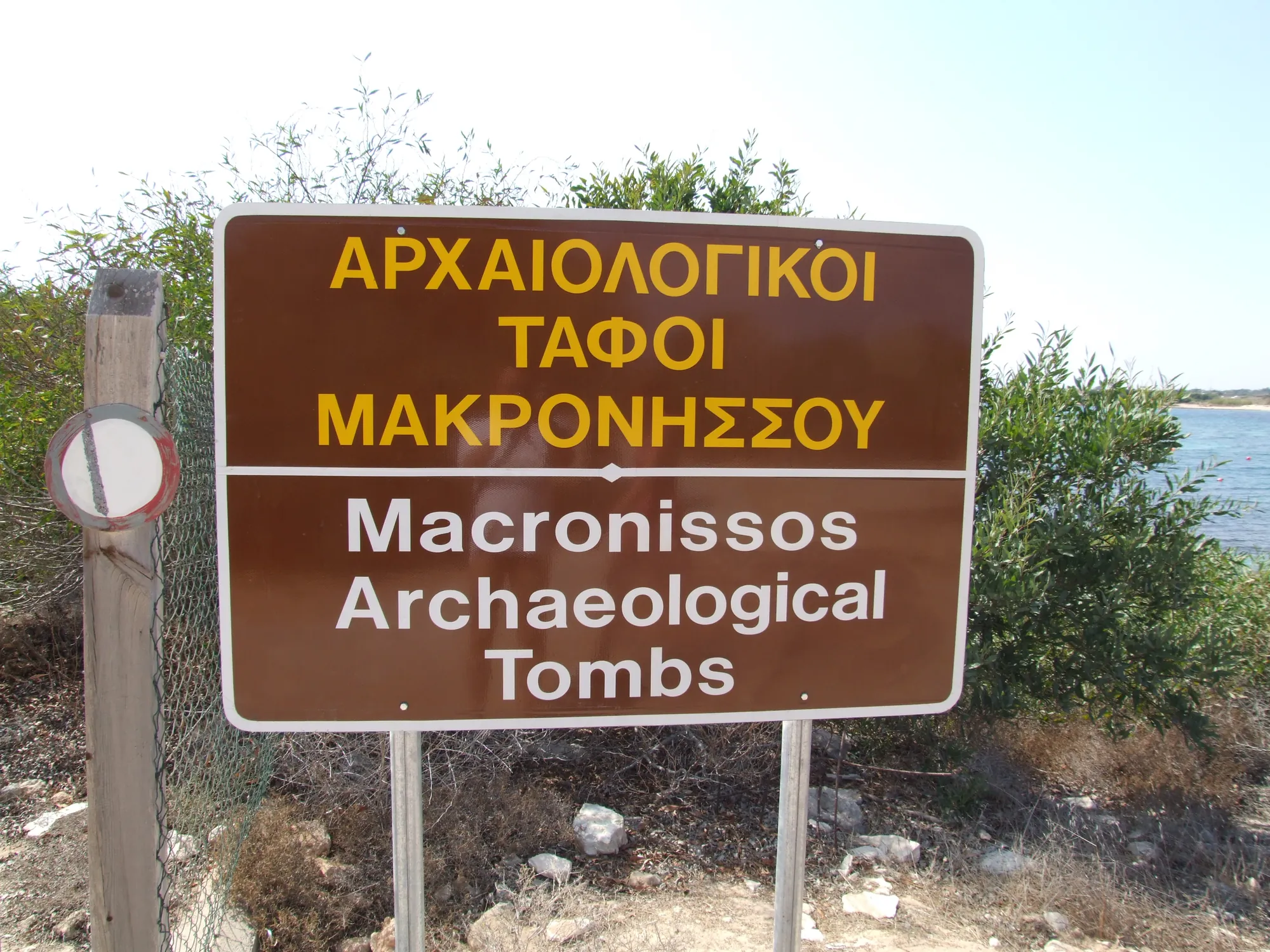 A brown sign reading Macronissos Archaeological Tombs in Greek and English, with bushes and sea behind