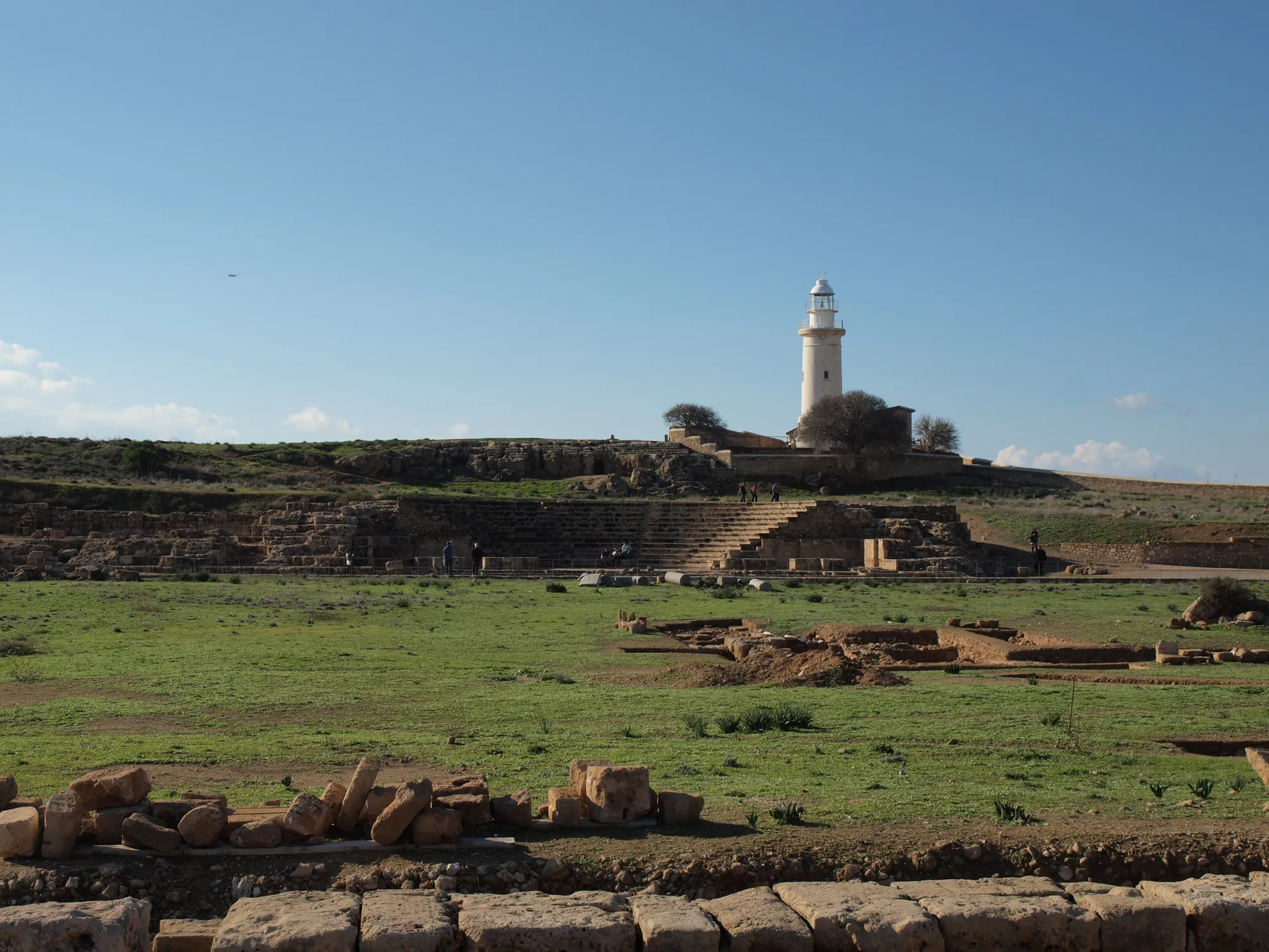 A grassy open area edged with low stone foundations, rising to the curved seating of a small theatre and a white lighthouse