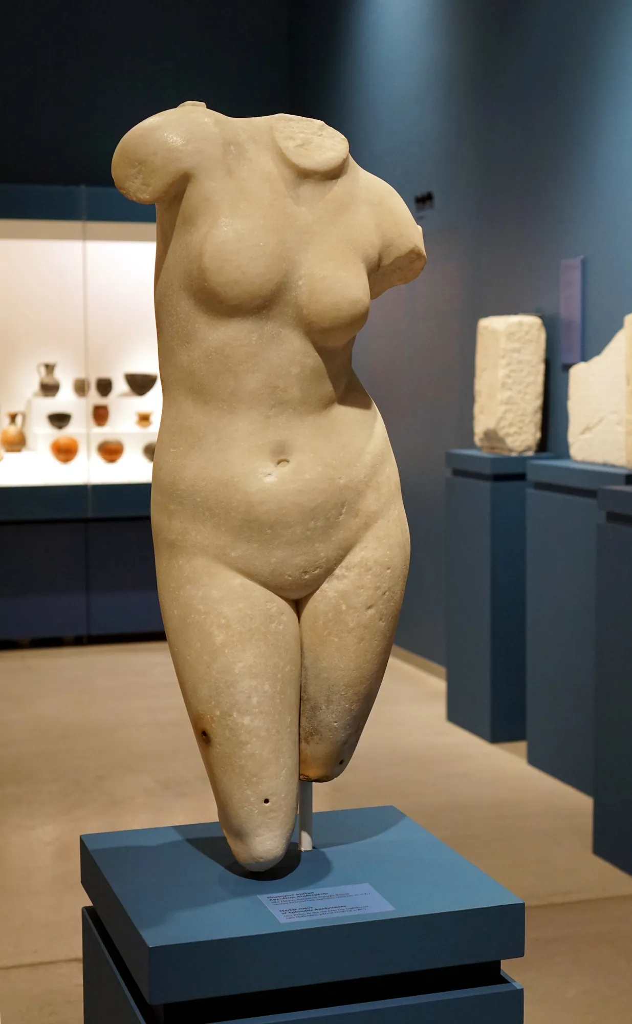 A headless marble statue of a nude female figure on a dark plinth in a museum gallery