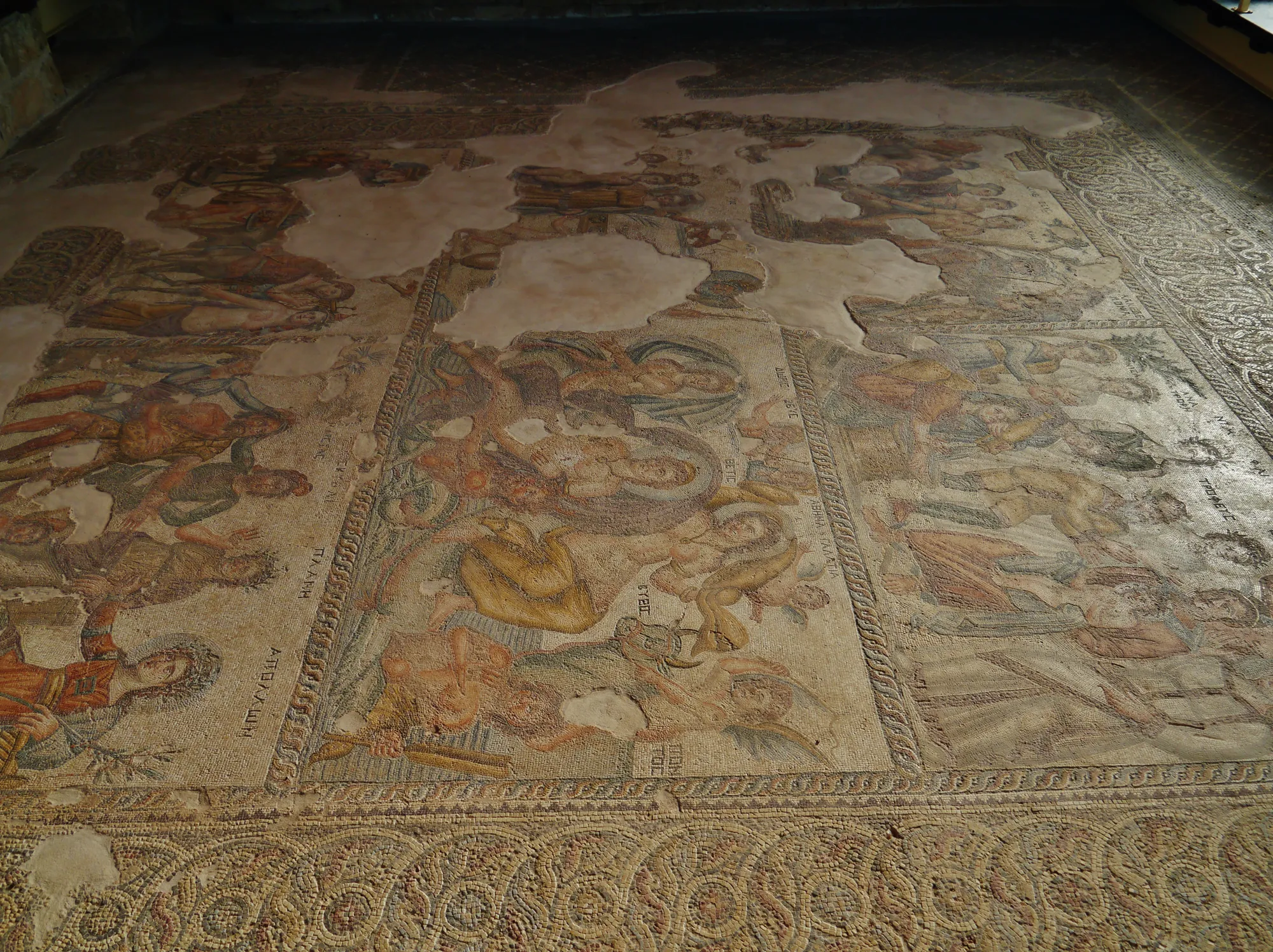 A large floor mosaic seen at an angle, divided into panels of figures, with patches of plain plaster where pieces are lost