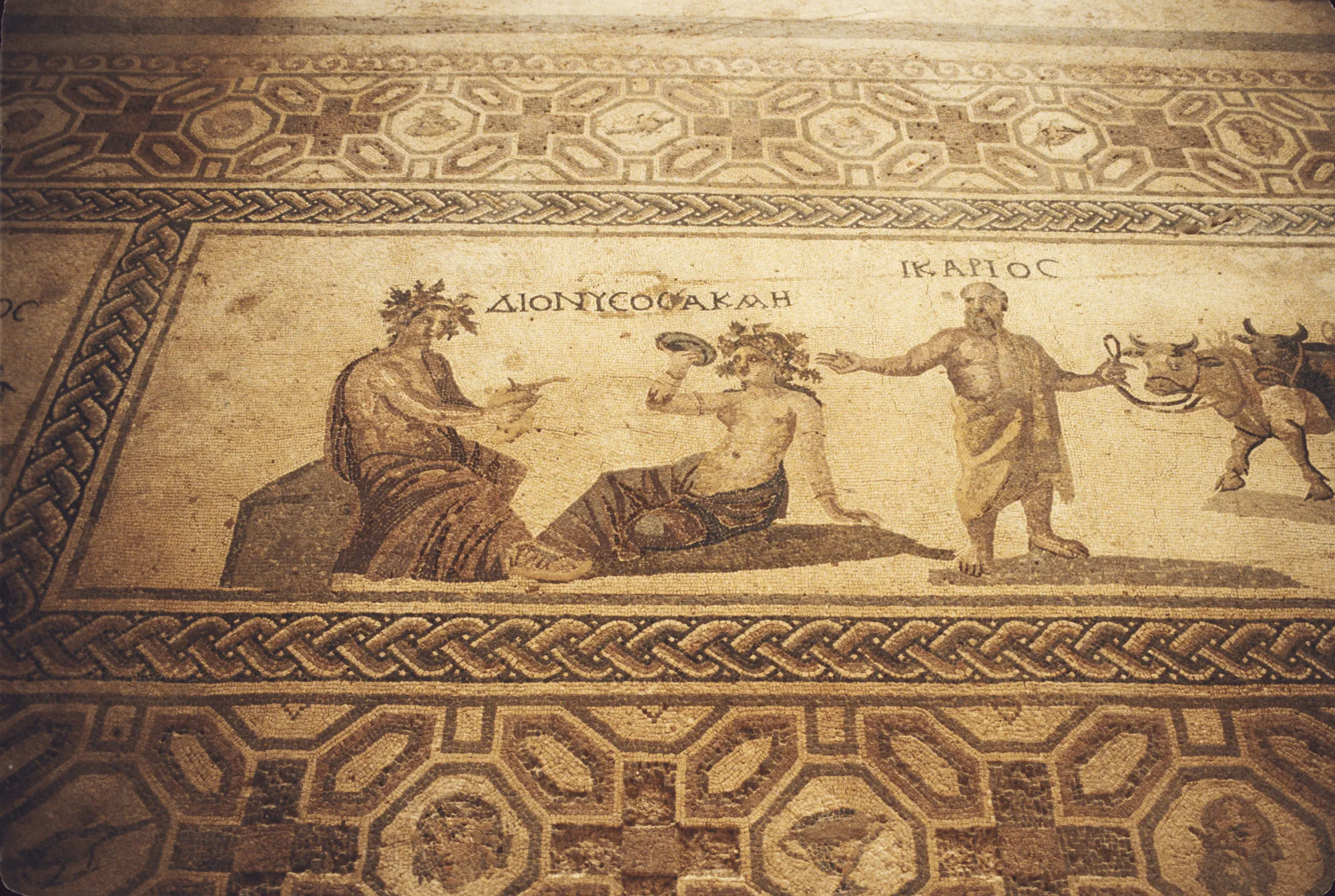A floor mosaic panel with Greek labels: a seated god, a reclining woman holding a cup, and a man leading oxen and a cart