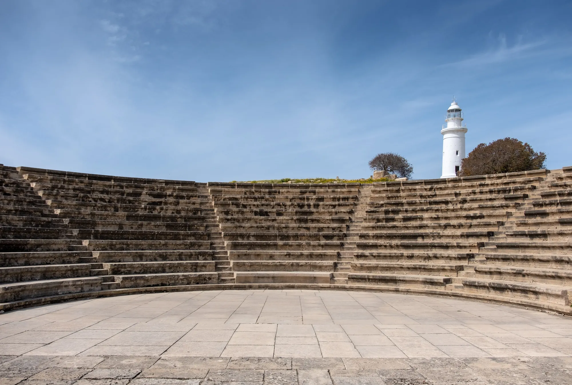 Rows of curved stone seats rising around a paved semicircular floor, with a white lighthouse above