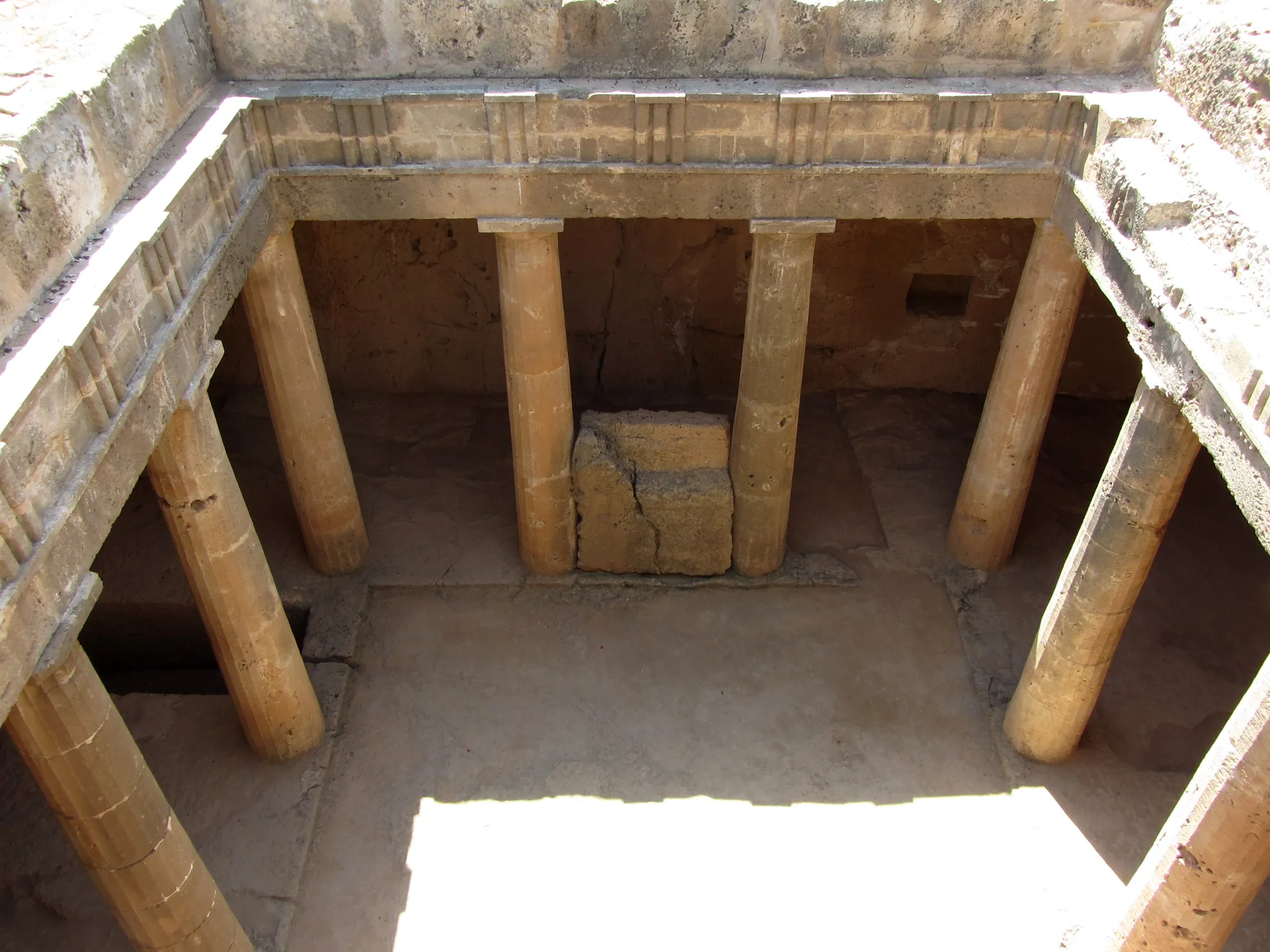 Looking down into a square court cut into rock, ringed by plain stone columns, with dark doorways behind them