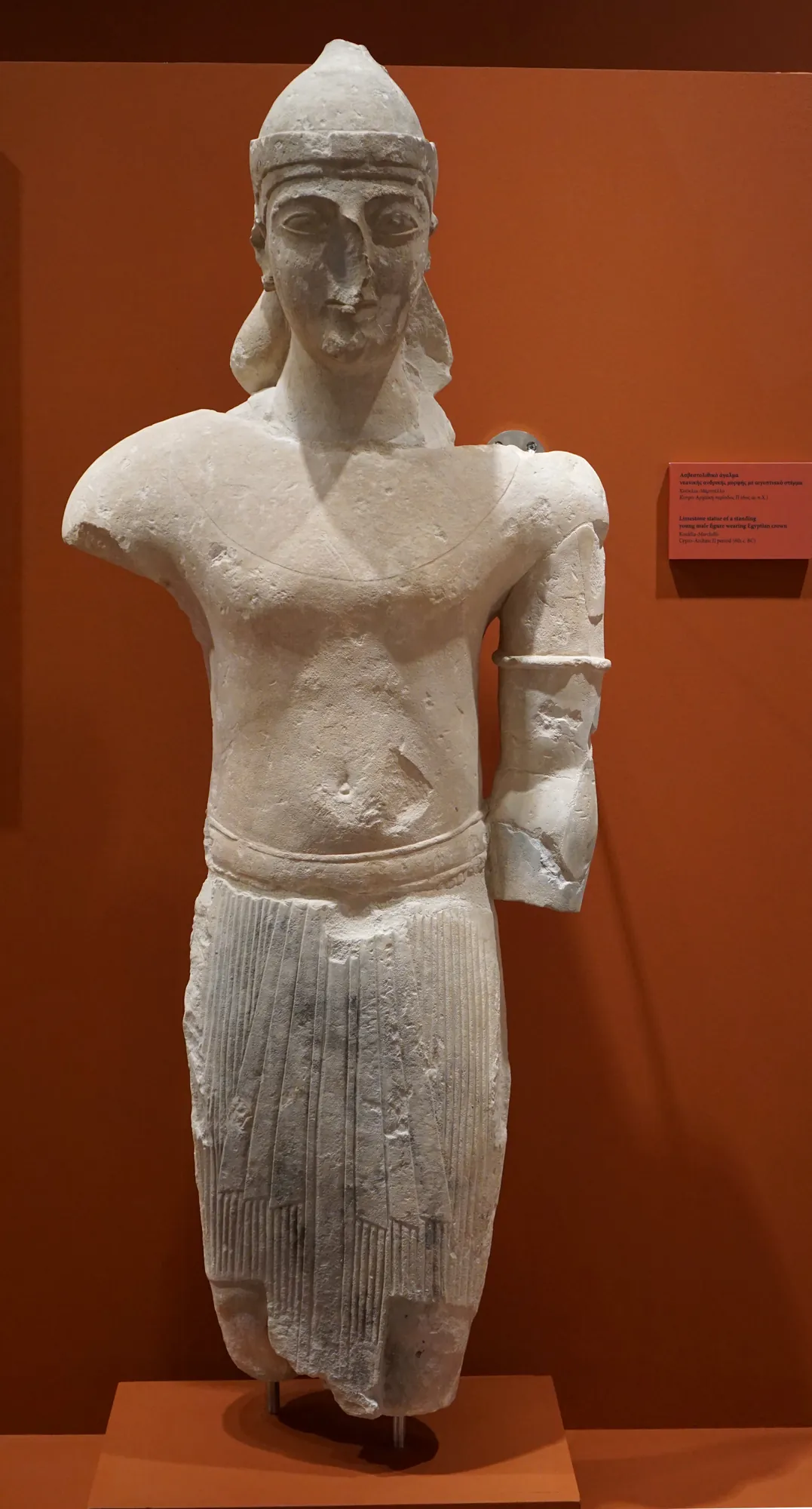Standing limestone male figure with a tall crown and kilt on a red wall