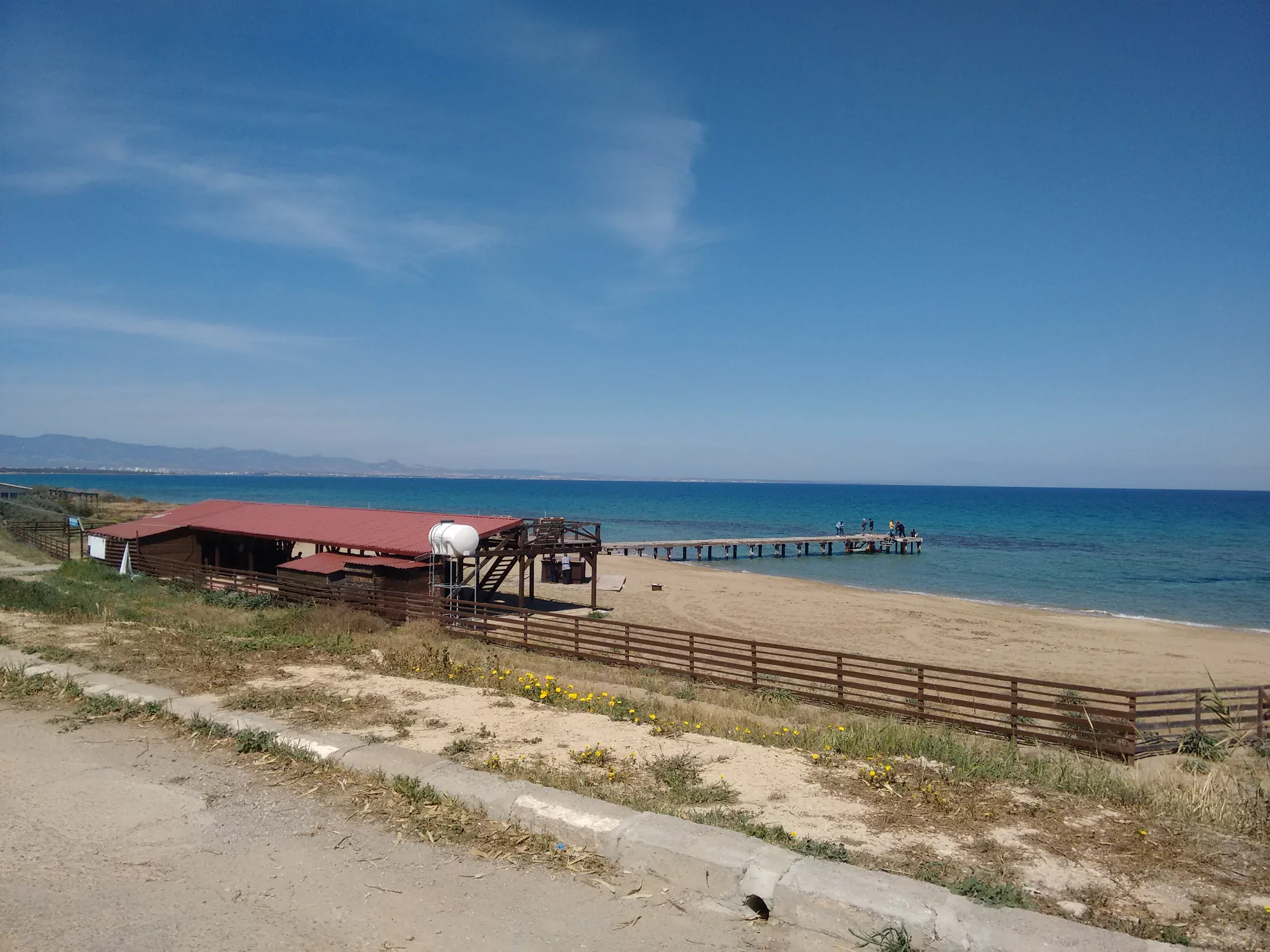 A sandy beach with a wooden pier and low scrub, the sea and blue sky beyond