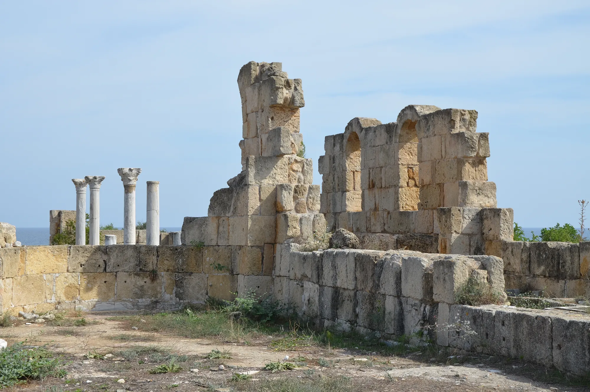 Low stone walls and column bases of a ruined basilica, with sky and sea haze behind