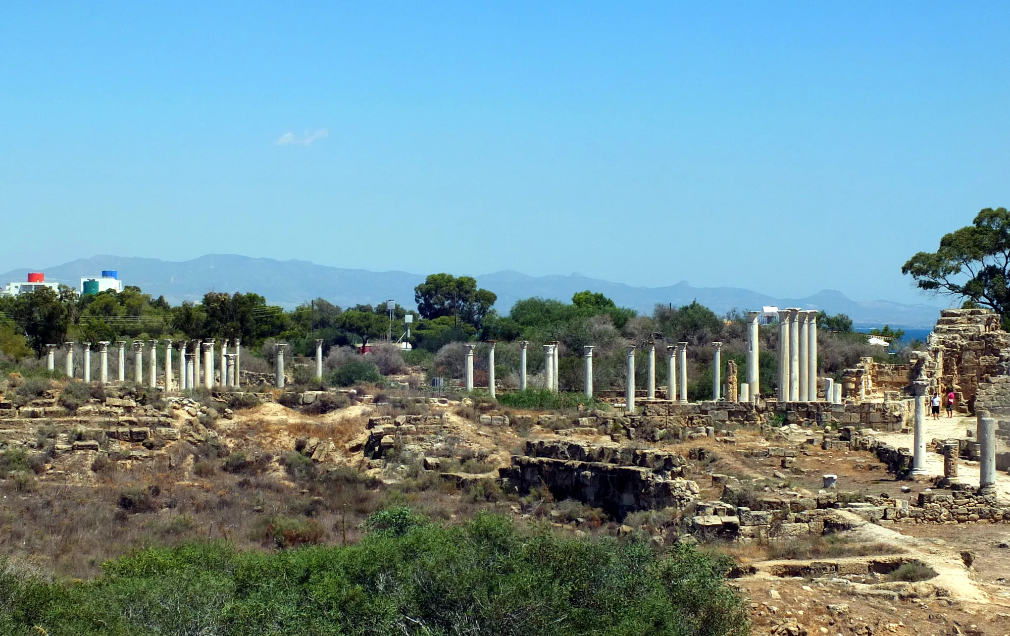 Distant white columns on a low ridge among scrub, with blue hills and sea beyond